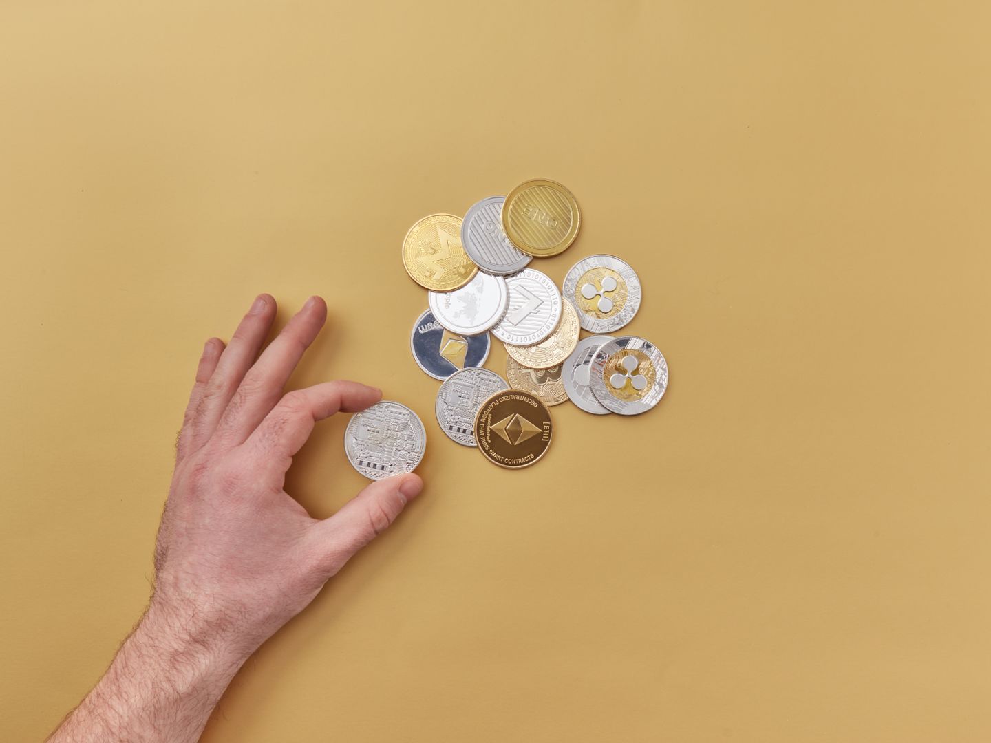 A hand picks cryptocurrencies from a mix of gold and silver coins on a vibrant yellow background.